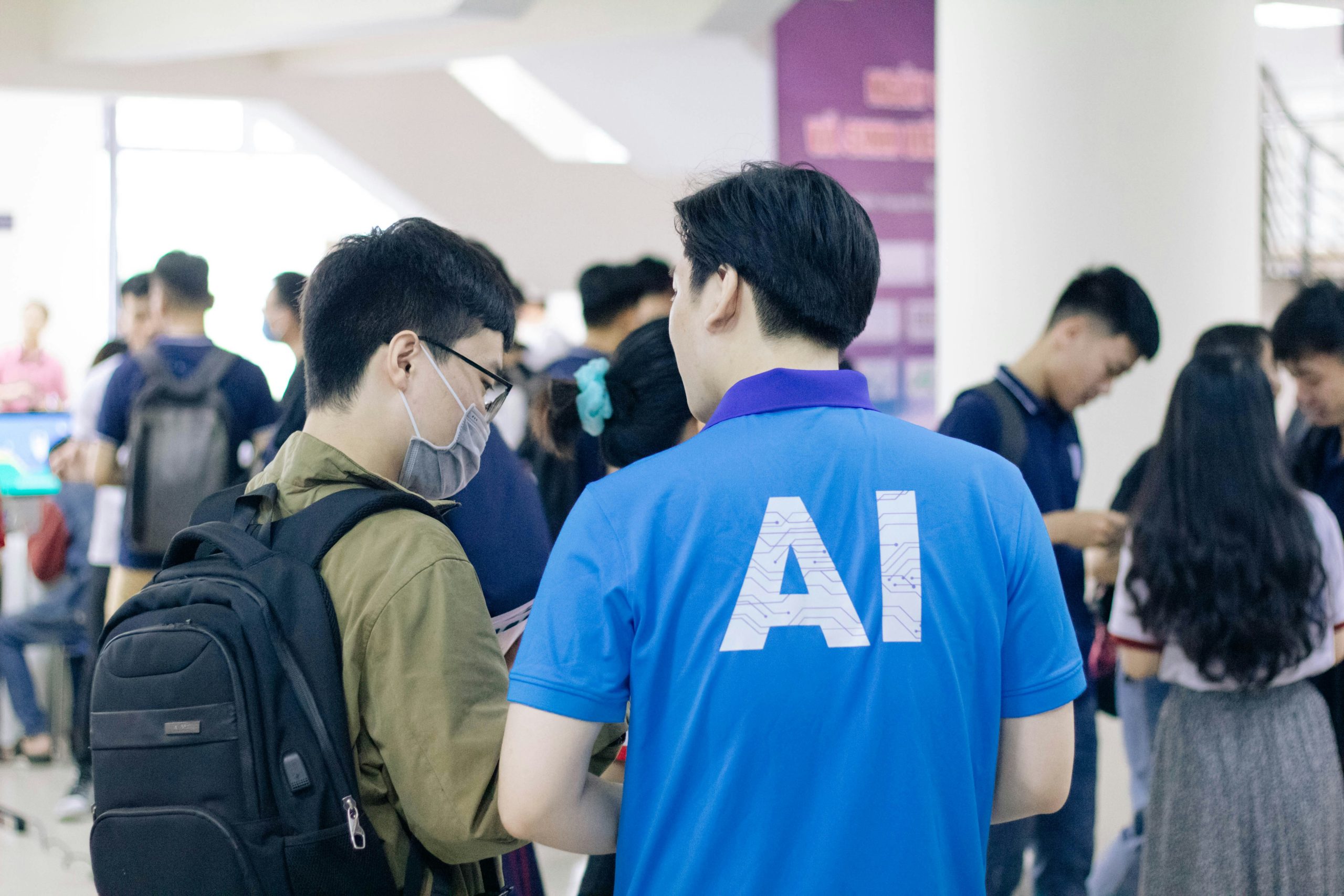 A person wearing a blue shirt with white AI text on the back stands near another person wearing a mask and backpack, in a cro